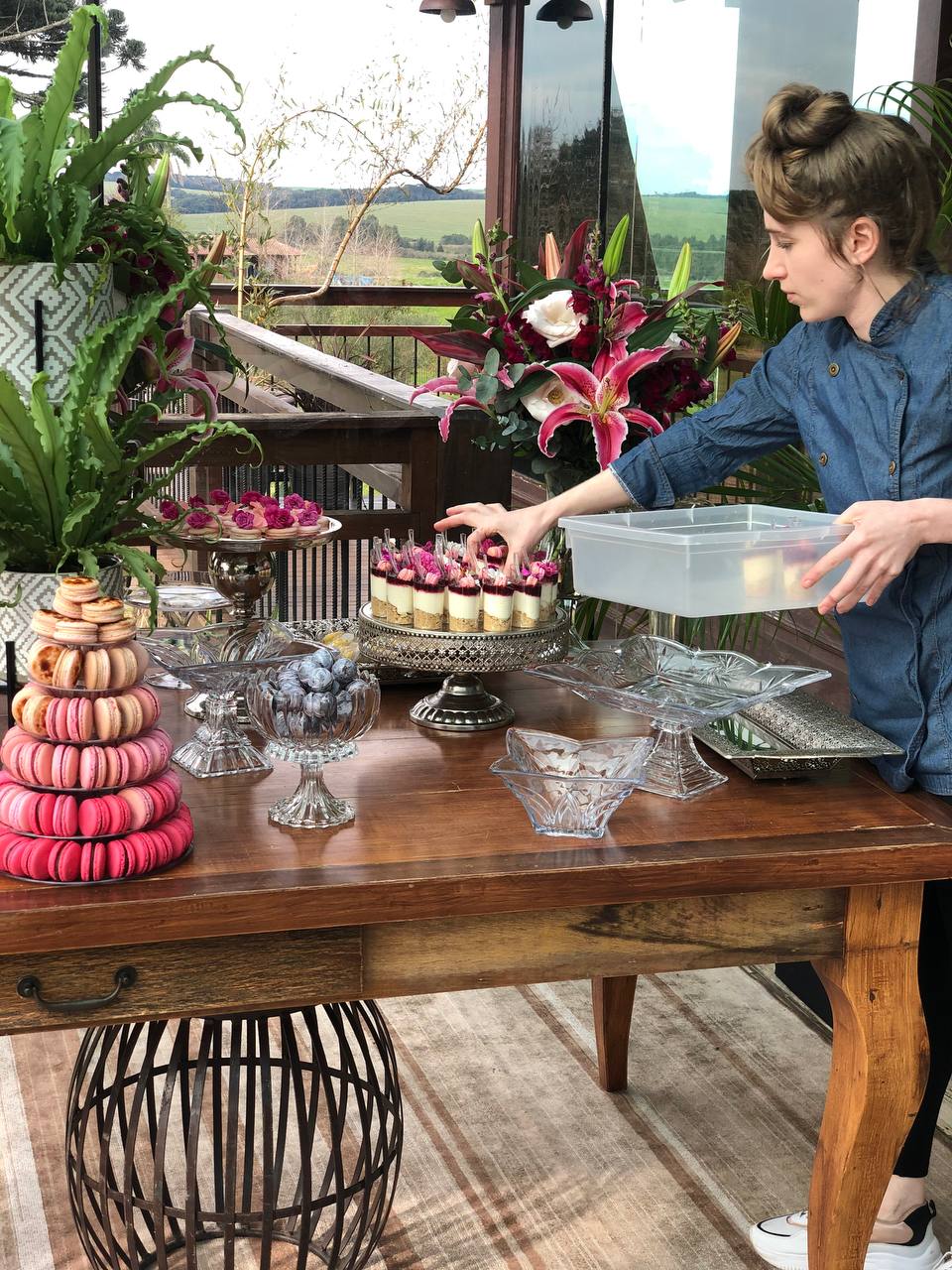 Mesa de doces sendo finalizada para um evento, com macarons, flores e taças de sobremesa